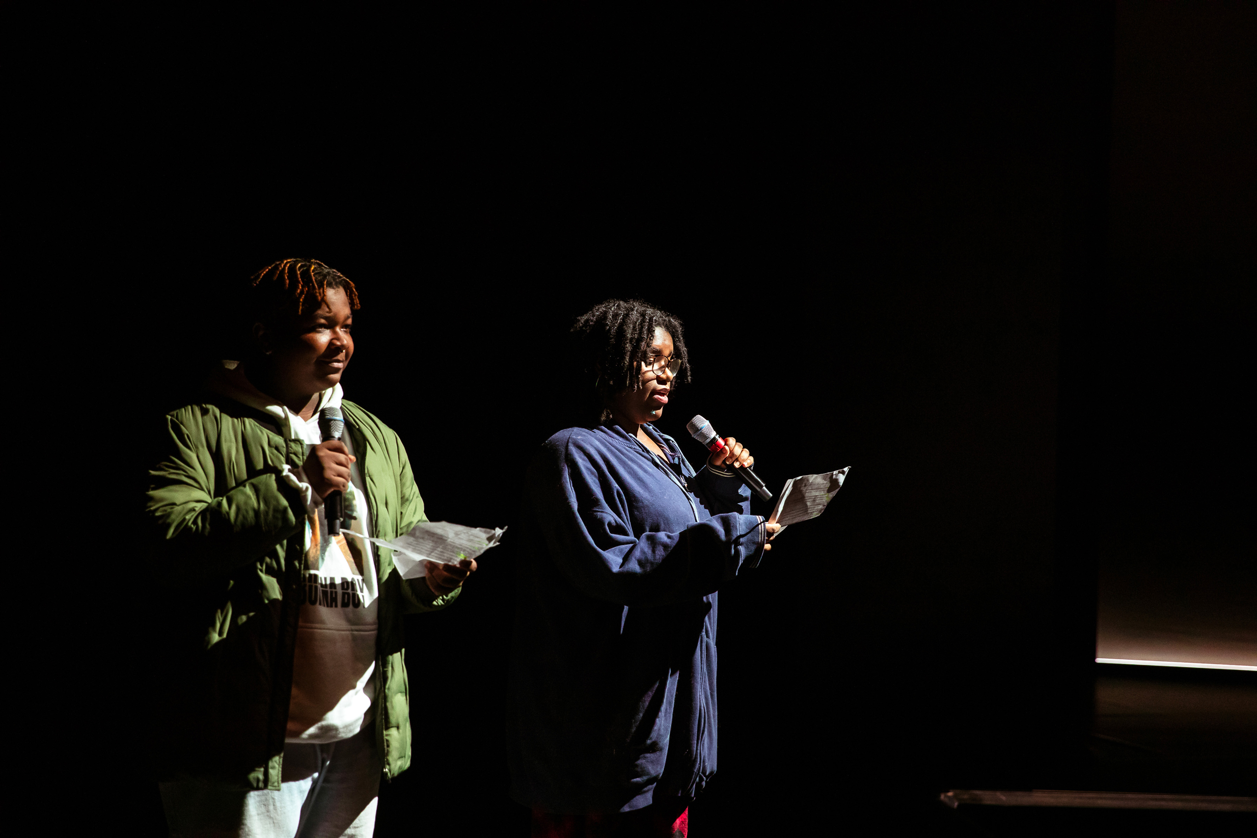 Two students in jackets stand on a darkened stage and read from a page into hand-held mics.