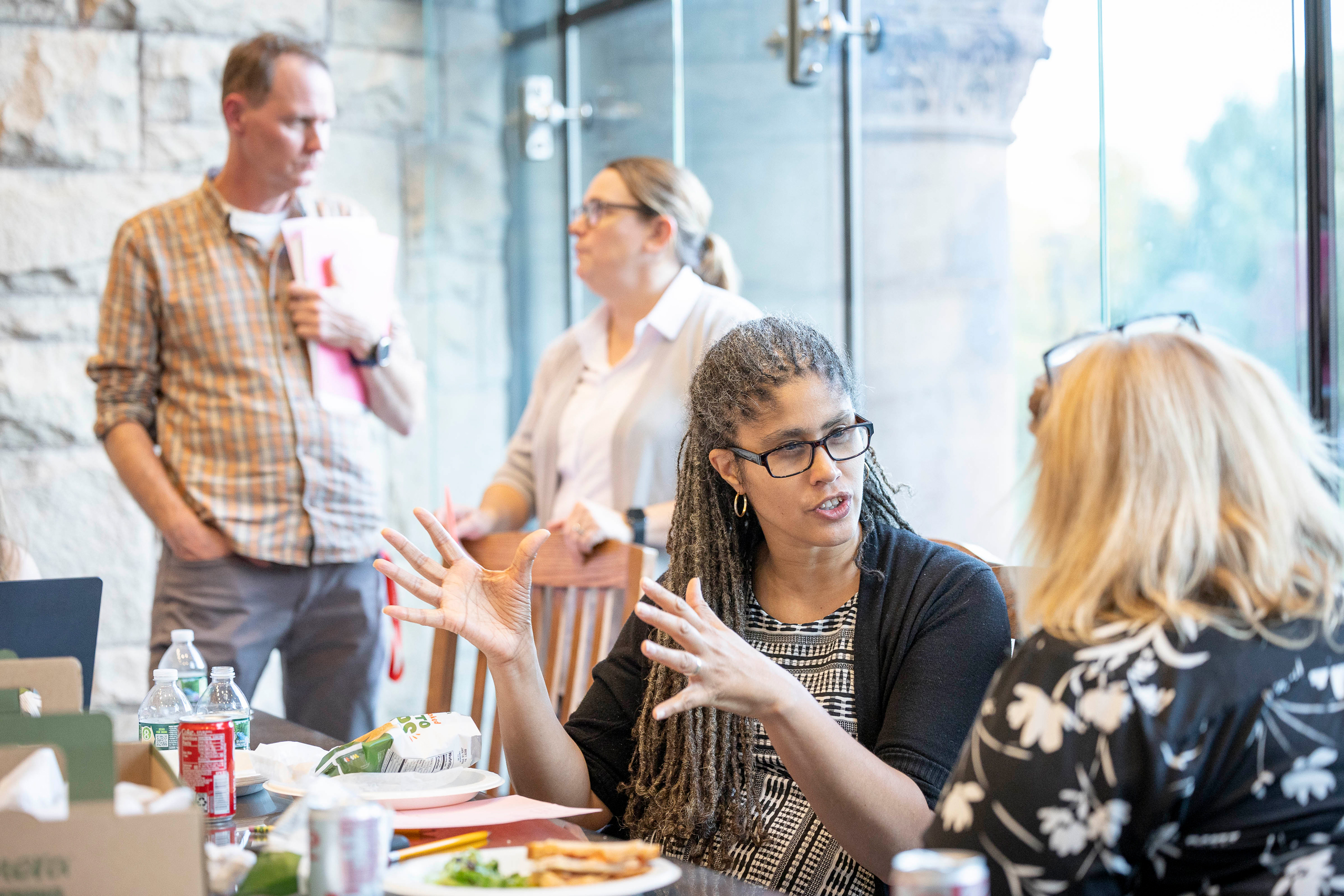 A teacher sitting at table in a glass-walled room gestures as she speaks with another teacher. Two more teachers stand behind them speaking.