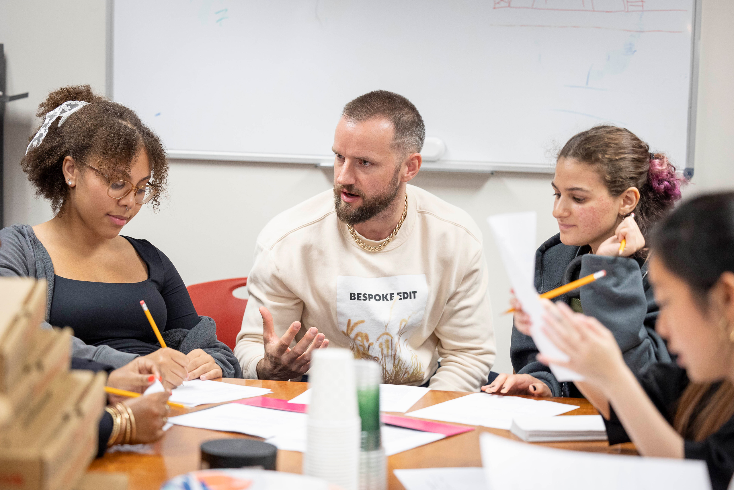 Actor Alex Ross in a sweatshirt and chain speaks to students sitting on either side of him taking notes.