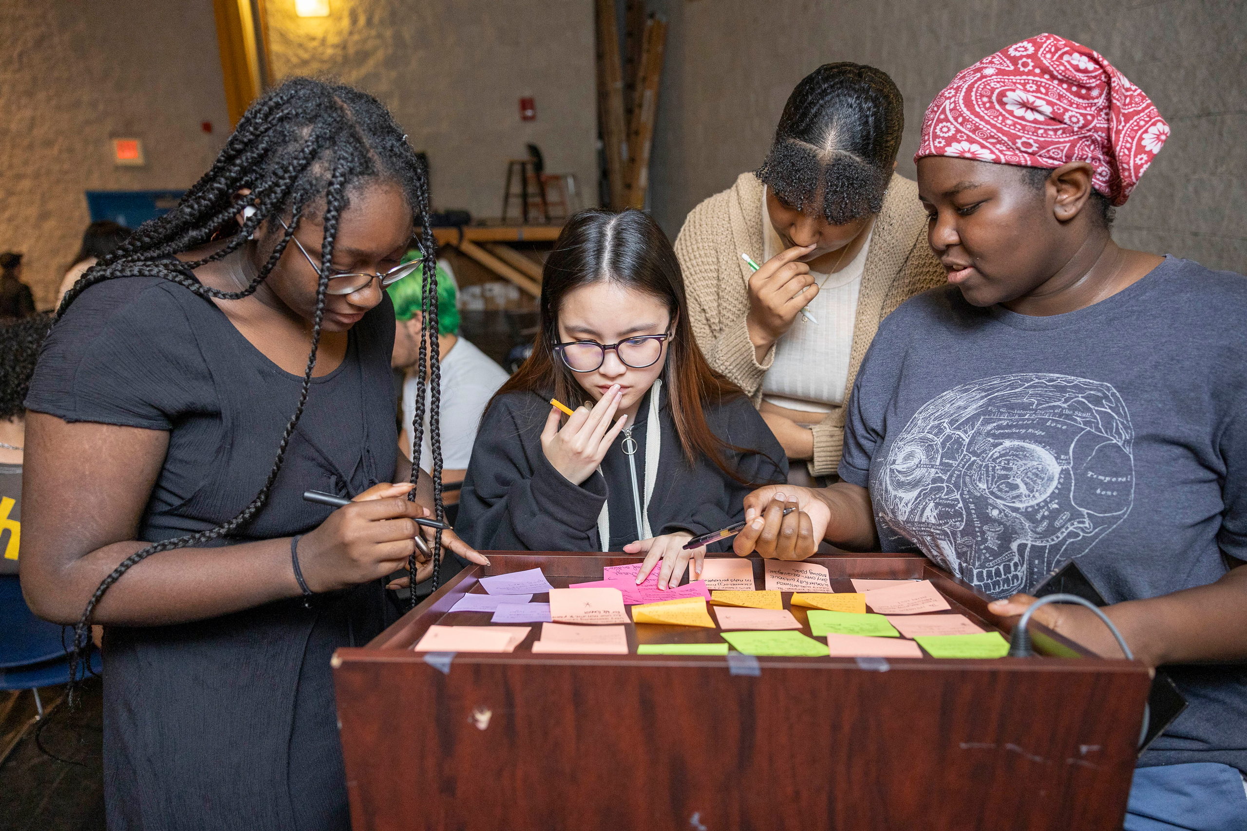 Four students in a dimly lit room gather around a podium with sticky notes spread across it.